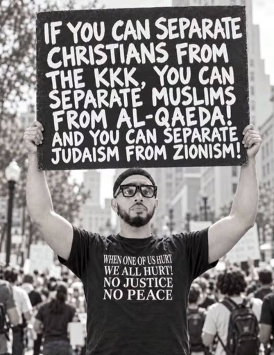 Photo of a bearded man at a demonstration with sunglasses and a shirt saying "When one of us hurts, we all hurt!. No Justice, No Peace". 

He is holding a sign aloft which reads:
 "If you can separate Christians from the KKK, you can separate Muslims from Al-Qaeda! And you can separate Judaism from Zionism!"